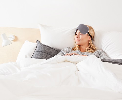 Woman tucked up in comfortable-looking bed wearing eye mask in grey